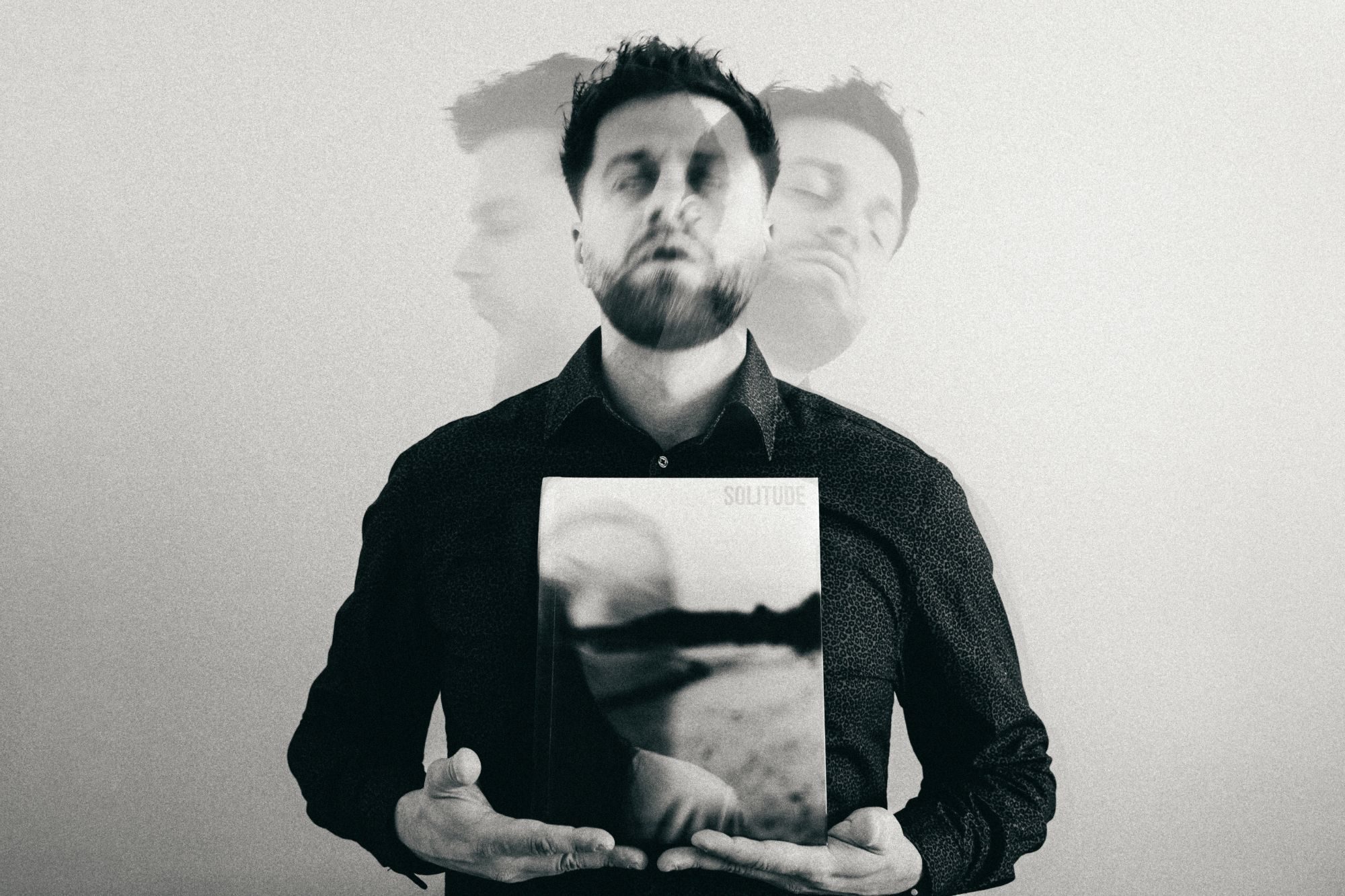 Dawid Kucięba trzyma album „Solitude.”; za nim prześwitują dwa odbicia jego twarzy. Czarno-białe.